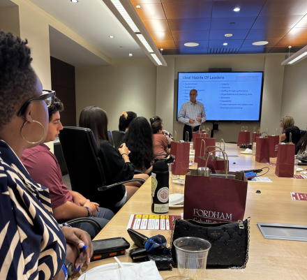 A speaker presents a slide titled “Ideal Habits of Leaders” to a group of students seated around a conference table at Fordham.